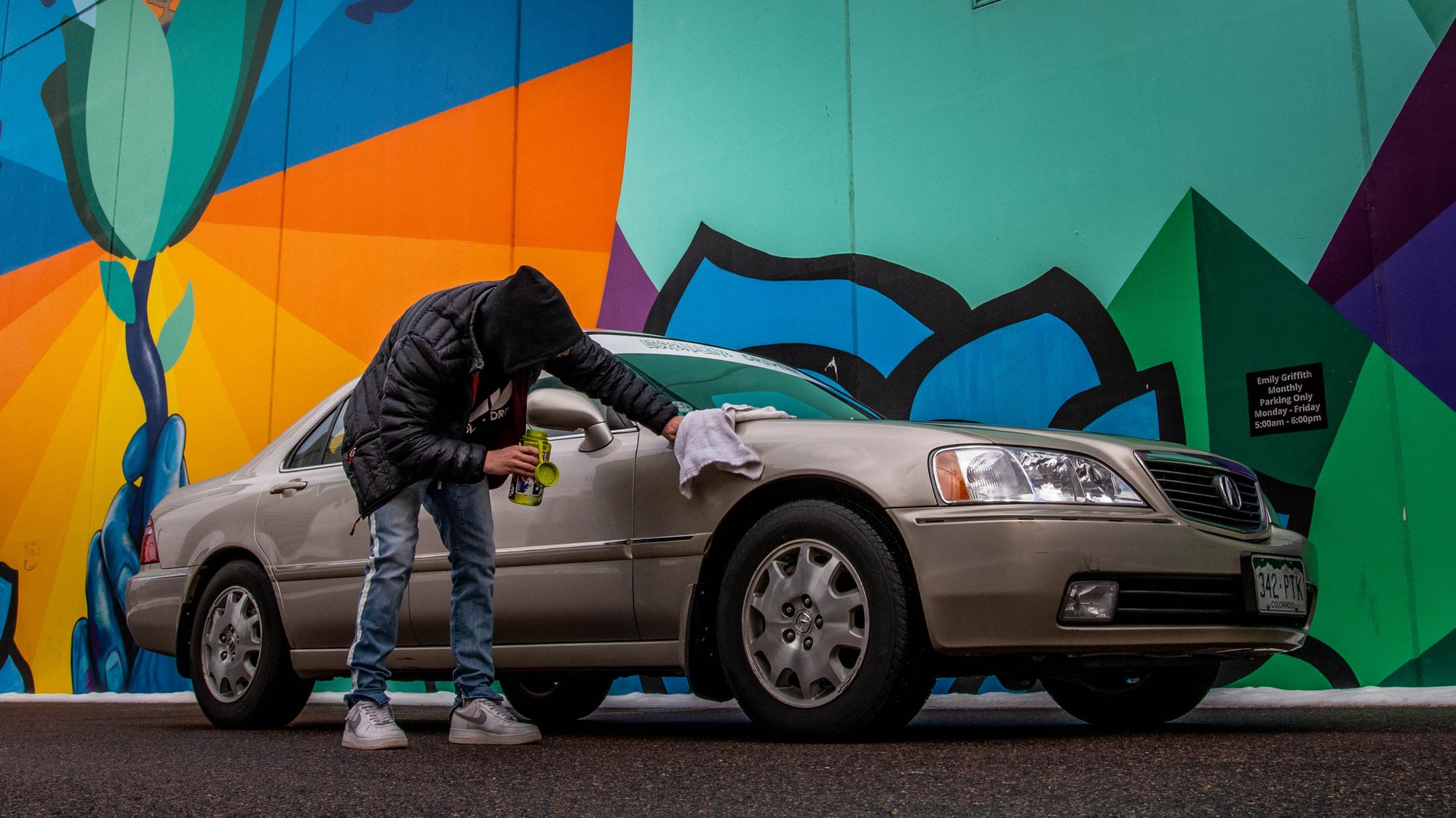 Hombre con capucha y chaqueta puliendo su auto frente a una pared colorida.